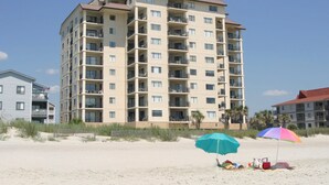 A multi-story hotel with balconies, located on a sandy beach with beach umbrellas and beach chairs.