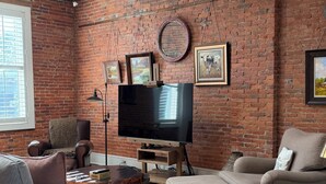 A living room with exposed brick walls, a flat-screen TV, leather armchairs, a patterned sofa, and a wooden coffee table. - Your Main Street Studio (Chagrin Falls)