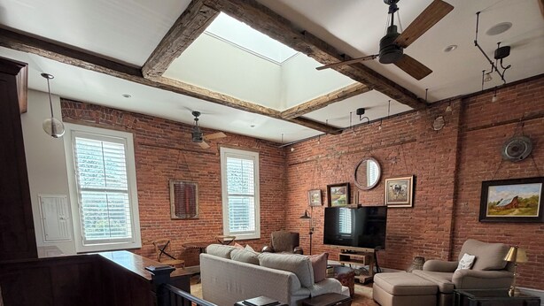 A spacious living room with exposed brick walls, a wooden ceiling, a large window, a ceiling fan, a fireplace, and a staircase. - Your Main Street Studio (Chagrin Falls)
