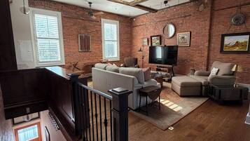 A spacious living room with exposed brick walls, a wooden ceiling, a large window, a ceiling fan, a fireplace, and a staircase.