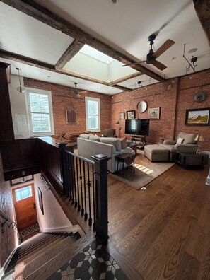 A spacious living room with exposed brick walls, a wooden ceiling, a large window, a ceiling fan, a fireplace, and a staircase.