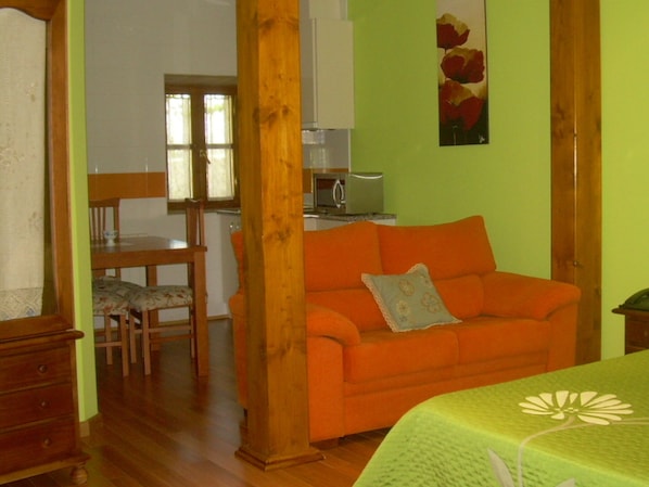 Une pièce avec un plancher en bois, une chaise en bois, un canapé orange orné d’un coussin à motifs floraux et un couvre-lit vert à motifs de fleurs blanches.