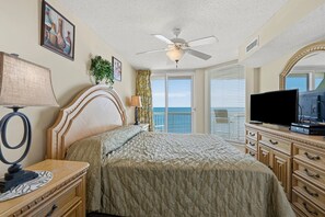 A hotel room with a bed, a television, a desk with a lamp, a ceiling fan, and a view of the ocean.