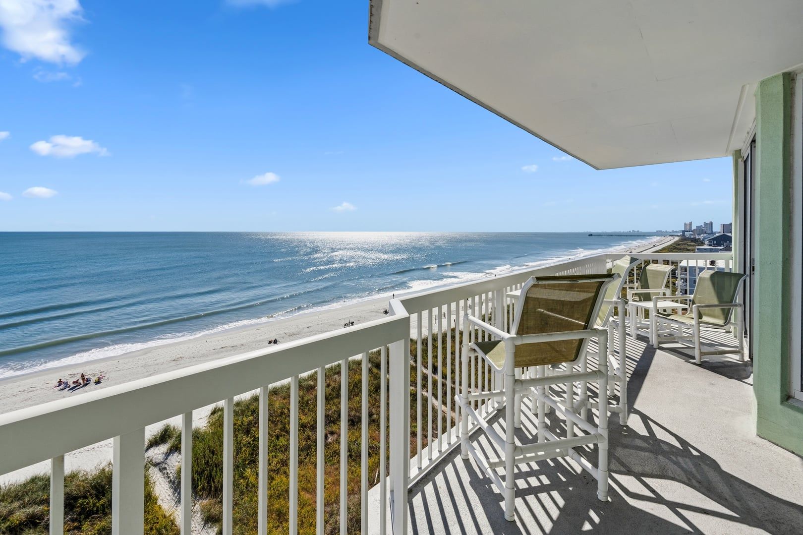A balcony with a view of the beach and ocean, featuring white railings and several chairs.