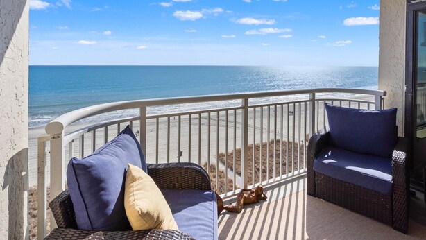 A balcony with wicker furniture and cushions, overlooking a beach and ocean.