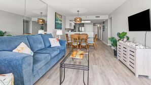 A modern living room with a blue sofa, a glass coffee table, and a dining area with wicker chairs.