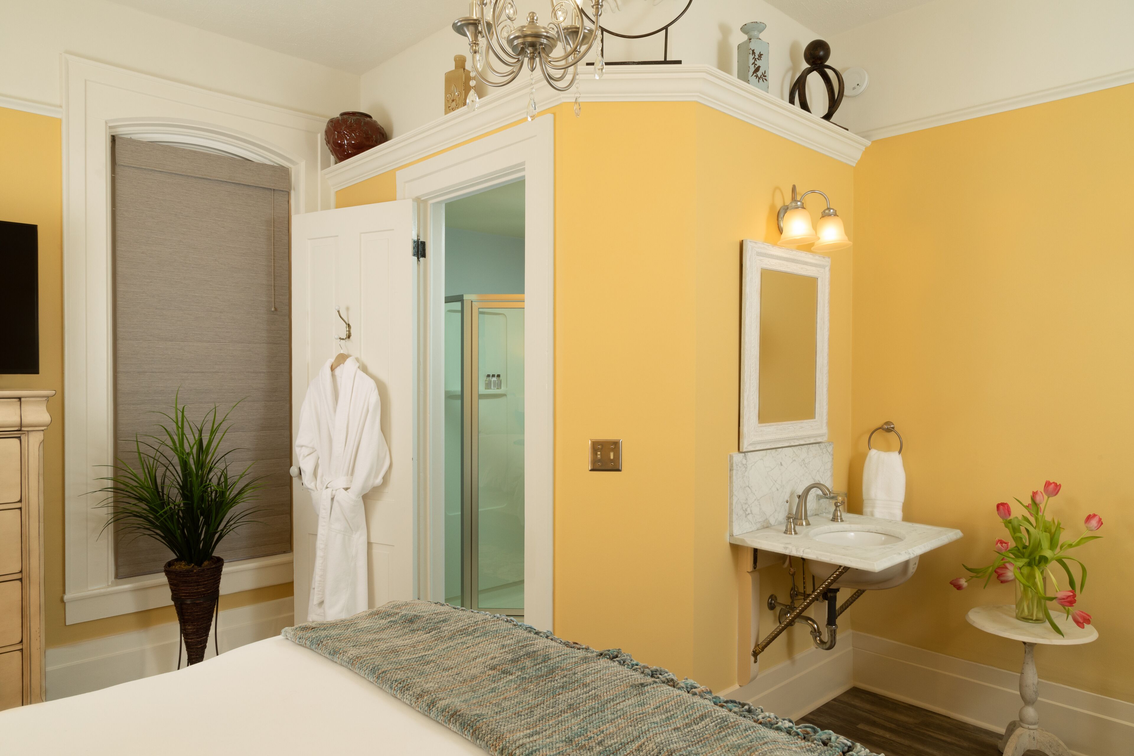 A bedroom with a bed, a bedside plant, a robe hanging on the door, a bathroom with a sink and mirror, and a small table with flowers.