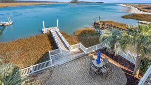 A wooden deck with a view of a calm body of water, a sandy beach, and a distant tree line. - Elegant luxury channel home w/plenty of relaxing space inside & out-deck, floating dock, grill (North Myrtle Beach)