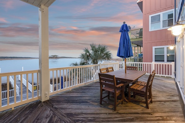 Um deck de madeira com mesa e cadeiras, com vista para a água ao pôr do sol.