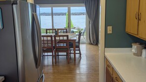 A kitchen with wooden cabinets, a stainless steel refrigerator, a dining area with a table and chairs, and a view of a body of water through large windows. - Beach Harbor Resort - Beach House Cottage (Sturgeon Bay)