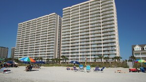 Ein Hotel direkt am Strand mit mehreren Etagen, Balkonen und Strandstühlen.
