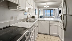 Una cucina moderna con piano cottura integrato, lavastoviglie e frigorifero. Dispone di mobili bianchi, un'ampia finestra con vista e un lampadario.