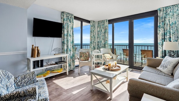 A spacious living room with a large TV, comfortable seating, a coffee table, and a view of the ocean.
