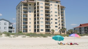 A multi-story hotel with balconies, located on a sandy beach with beach umbrellas and beach chairs.
