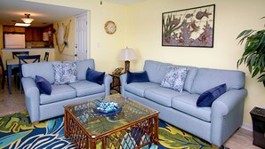 A living room with two sofas, a glass coffee table, a patterned rug, a clock, and a wall art piece.