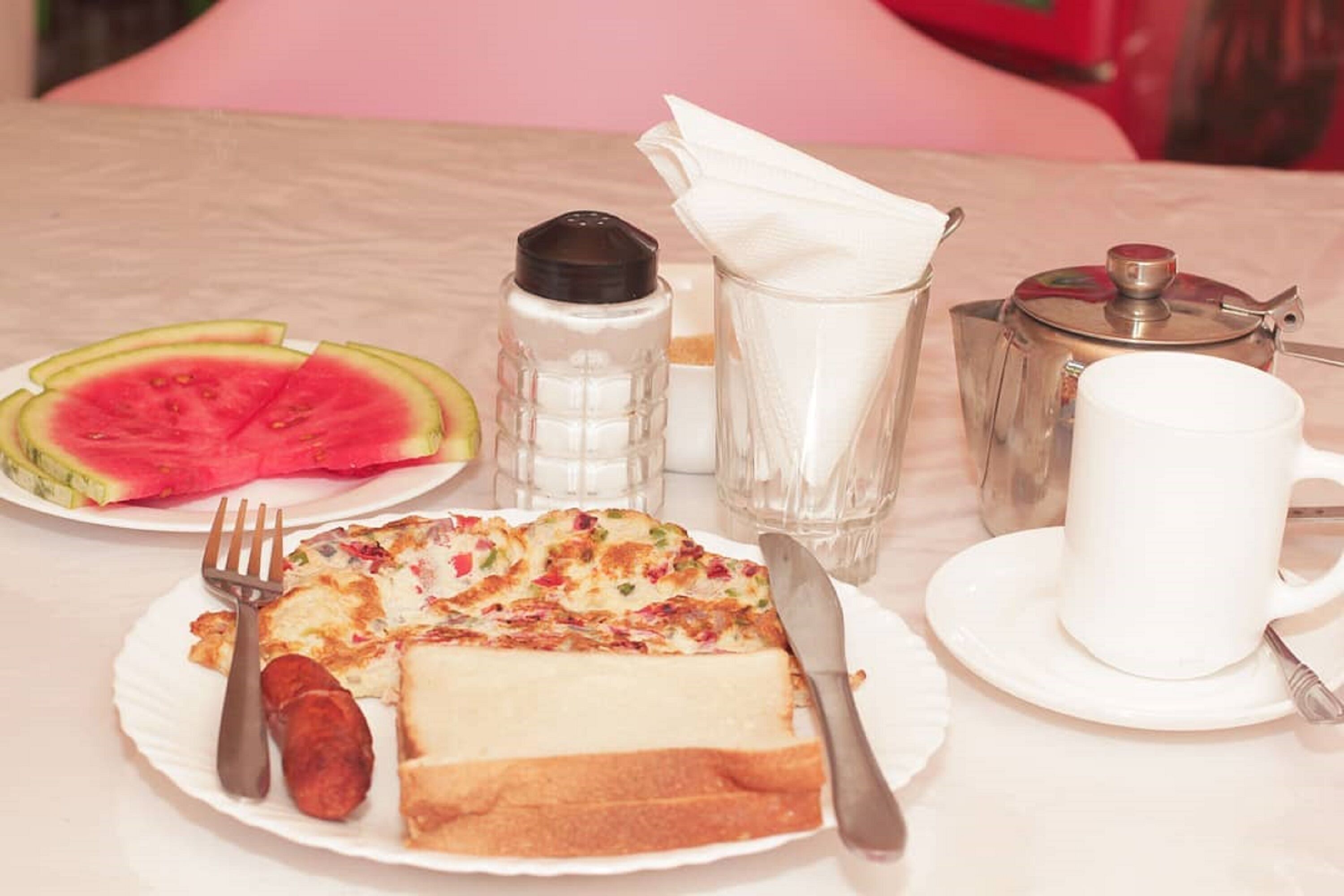 A breakfast setting with a plate of food, a slice of watermelon, a salt shaker, a pepper shaker, a coffee pot, and a cup.