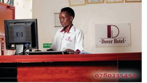 A hotel reception desk with a computer monitor, a sign for Dover Hotel, and a nameplate for the receptionist. - Dover Hotel Bondo (Bondo)