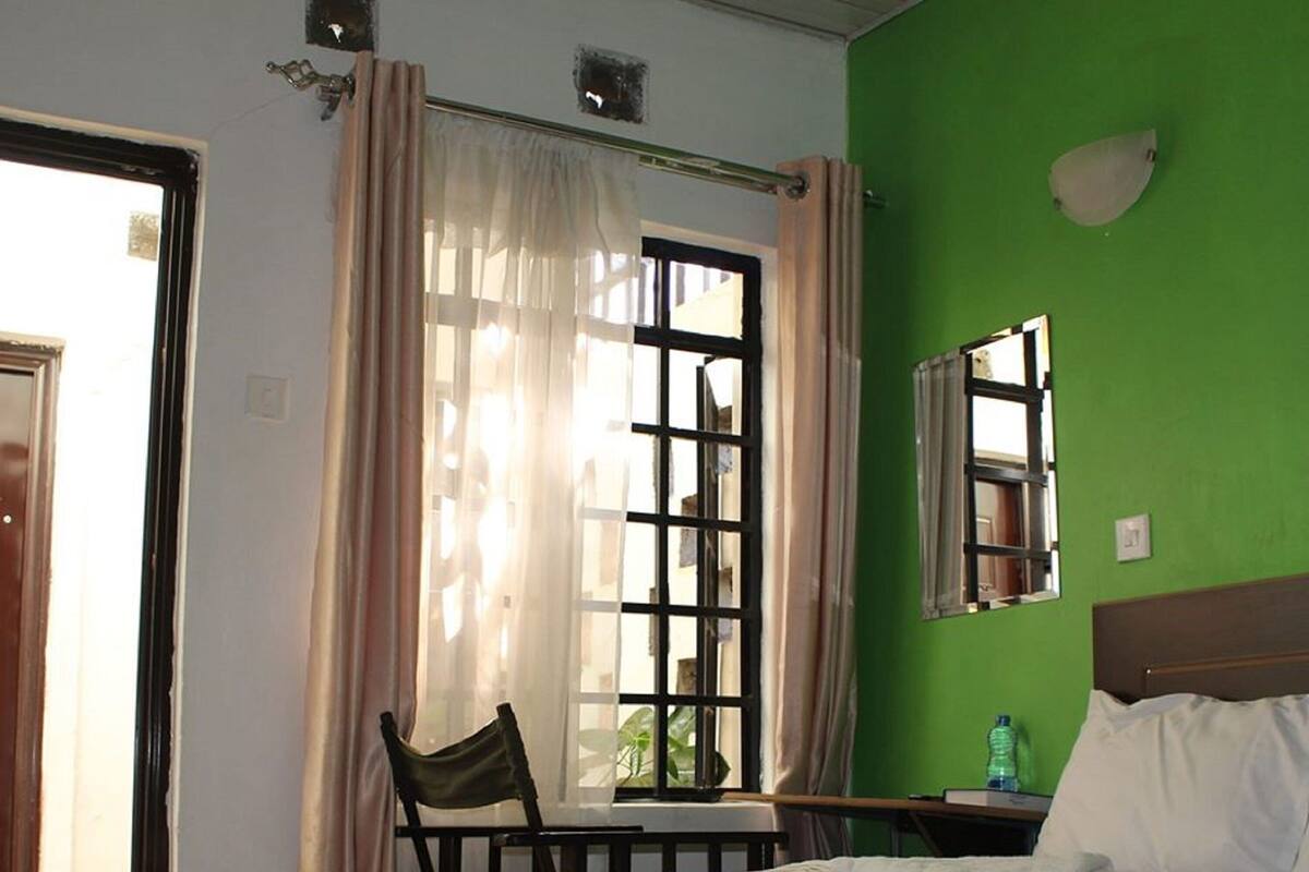 Un dormitorio con cama, una silla, una mesa, un ventanal con cortinas y una pared verde.