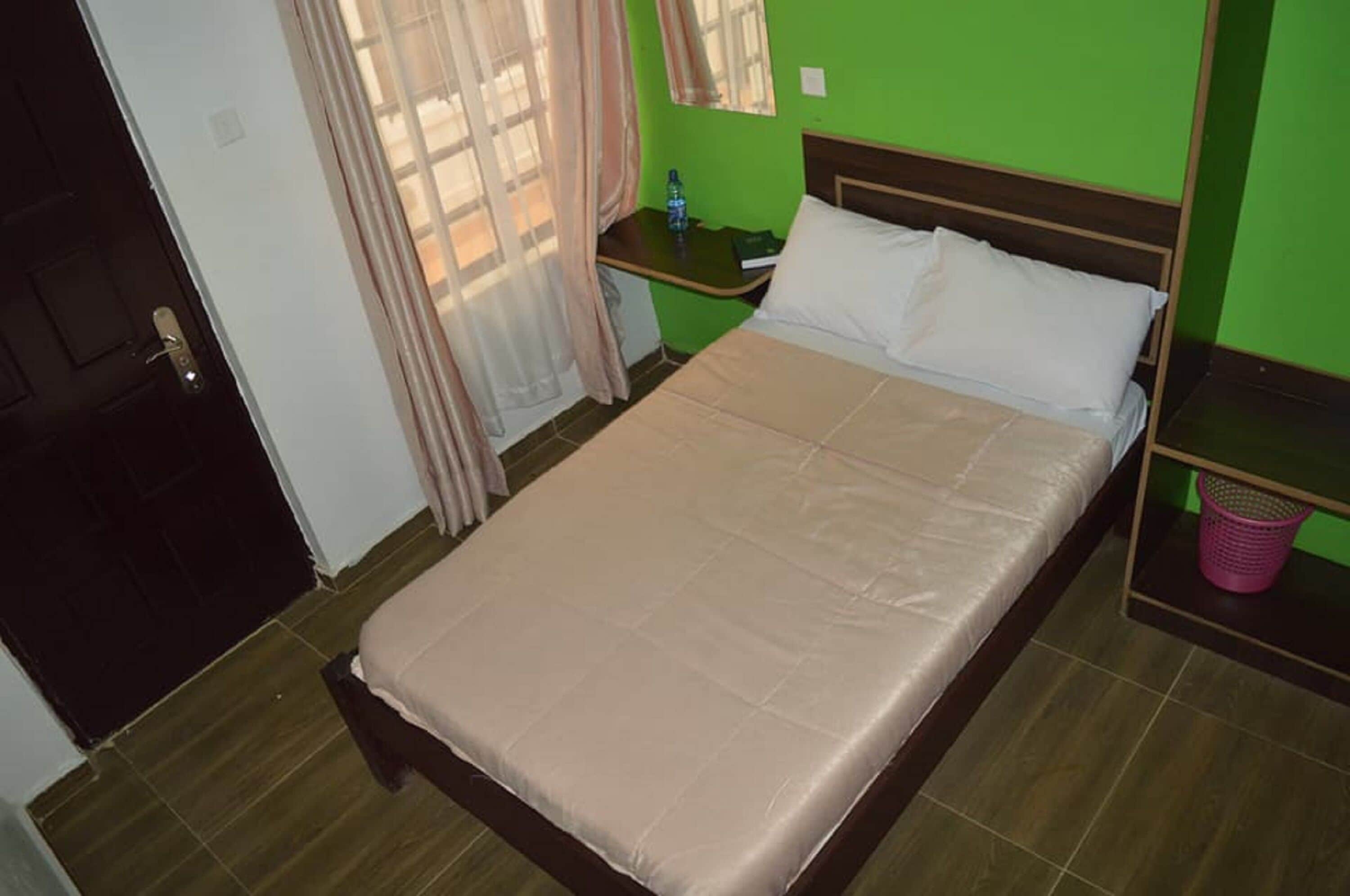 Una cama individual con colchón color beige y almohadas blancas en una habitación con paredes verdes y piso de madera.