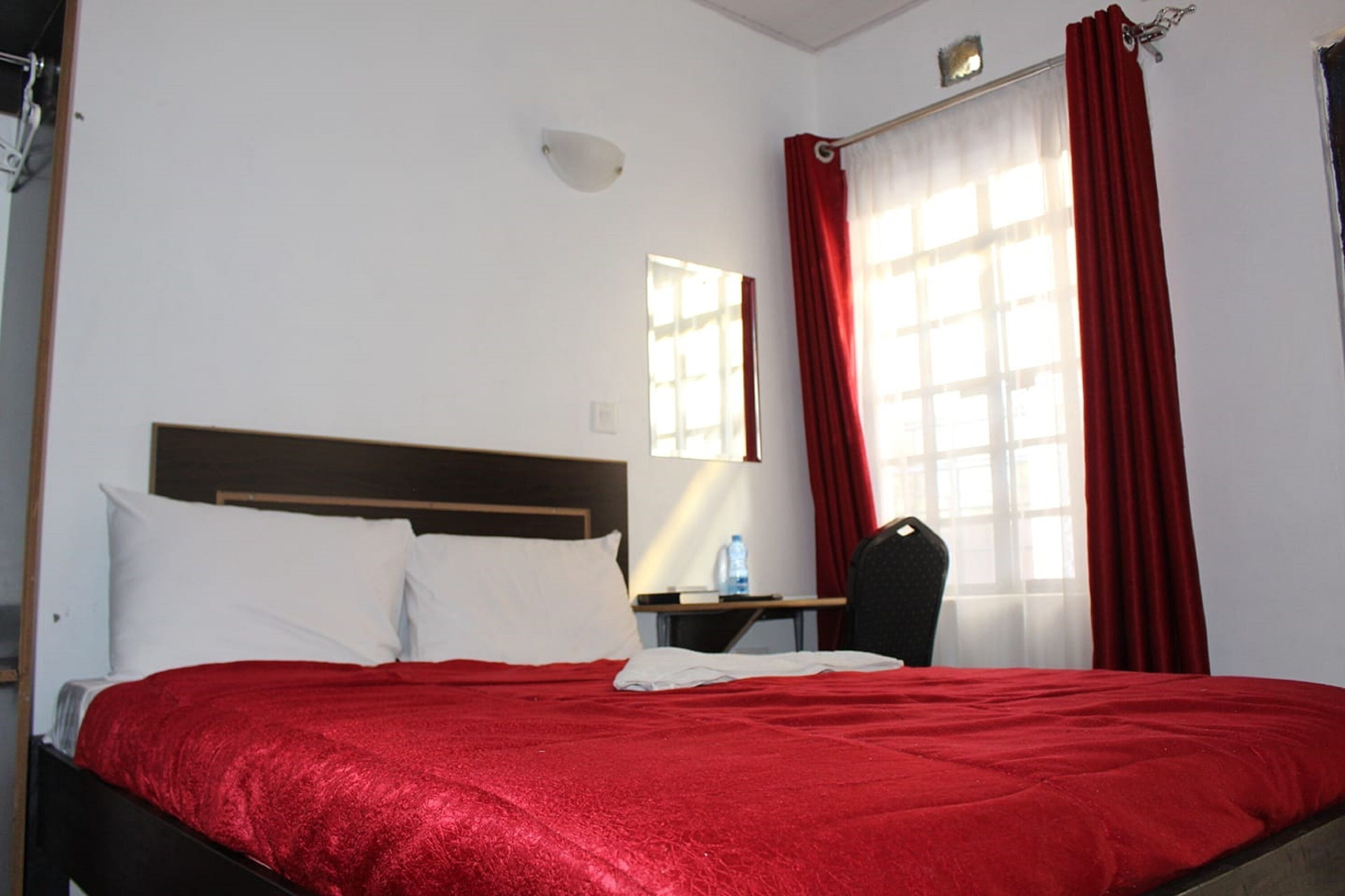 Una habitación de hotel con una cama, un escritorio, una silla, un ventanal con cortinas rojas y una lámpara fijada a la pared.