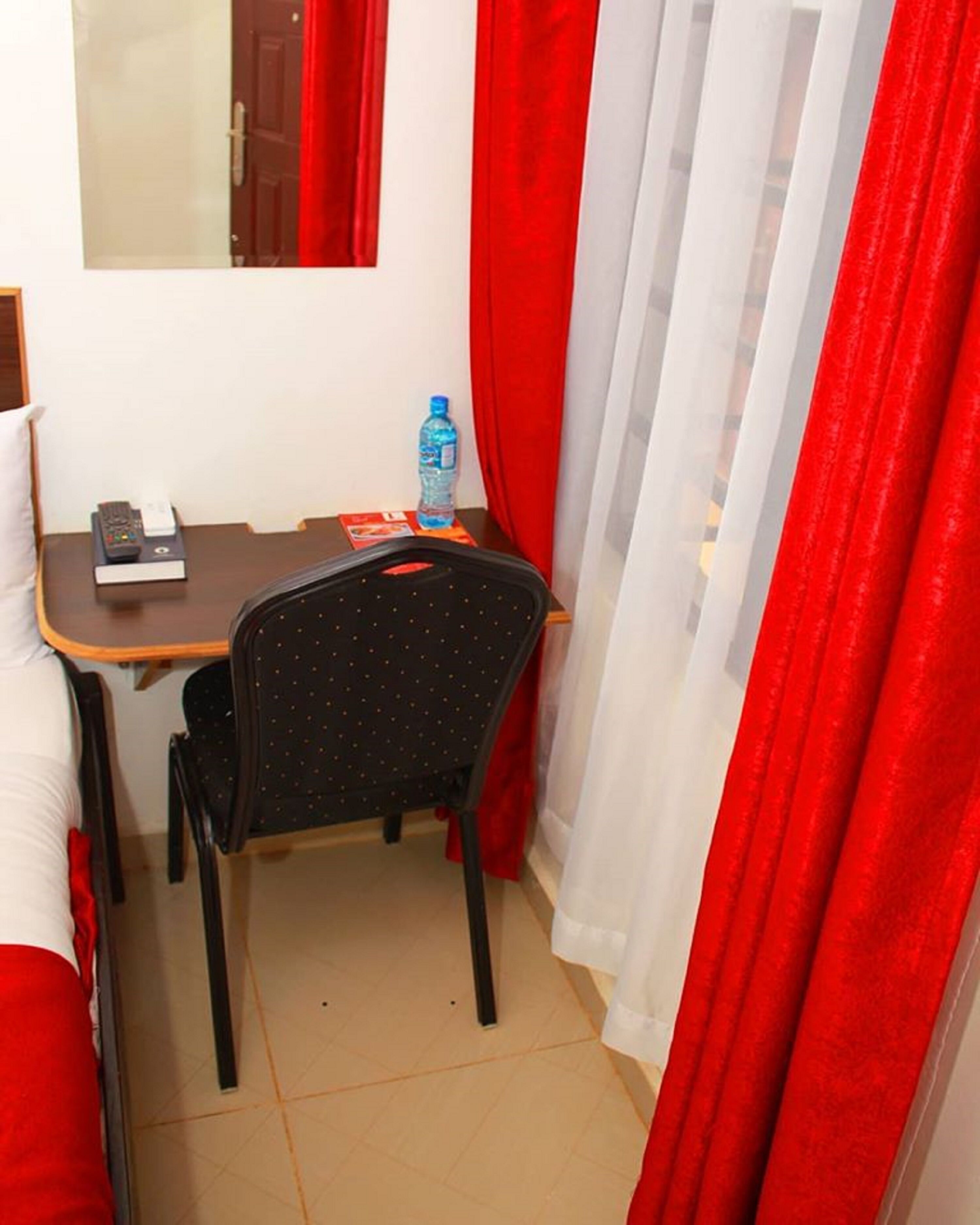 Una habitación pequeña con una cama, un escritorio, una silla, un ventanal con cortinas rojas y una botella de agua sobre el escritorio.