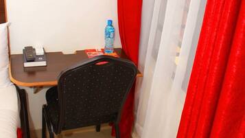 Una habitación pequeña con una cama, un escritorio, una silla, un ventanal con cortinas rojas y una botella de agua sobre el escritorio.