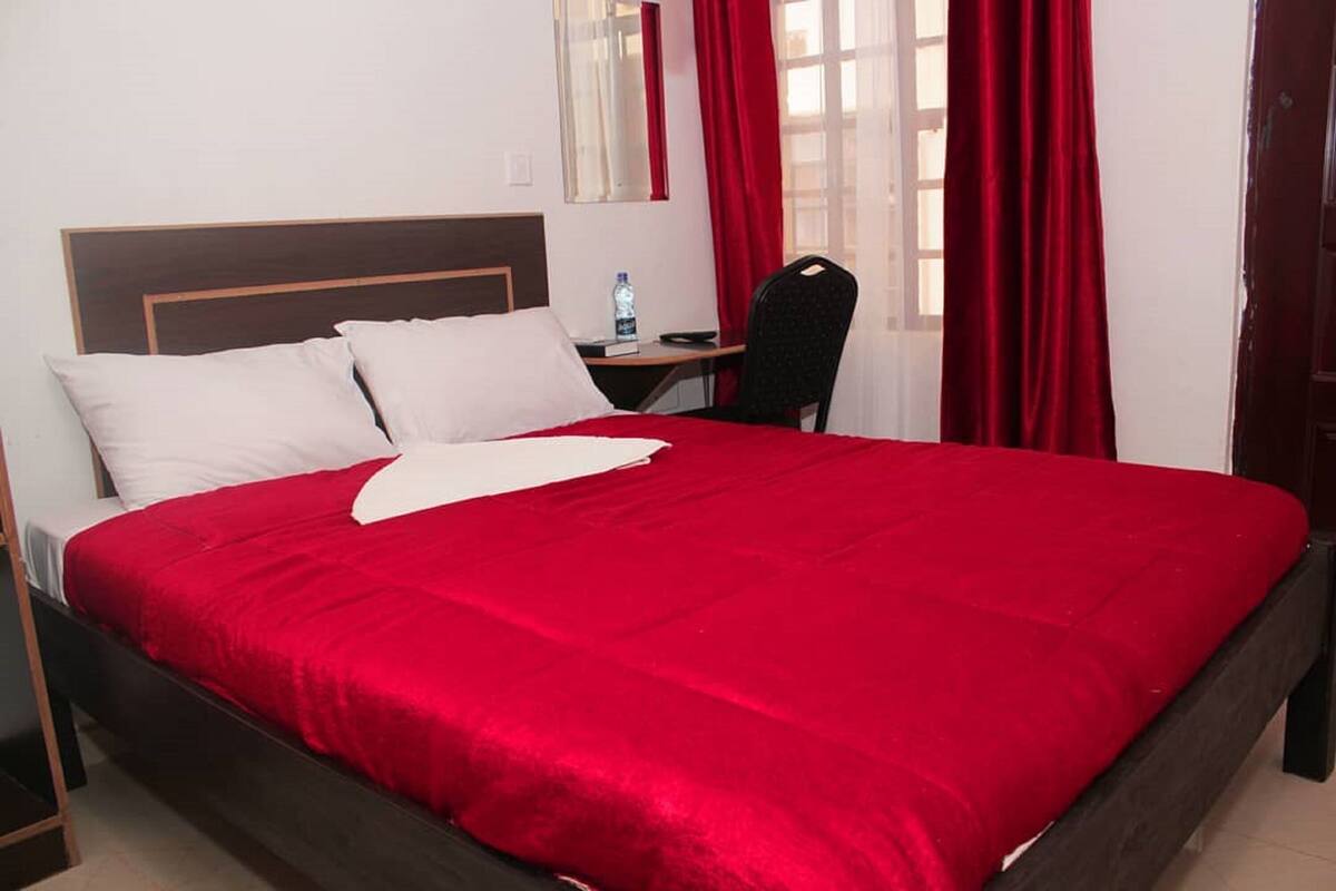 Una cama con una colcha roja, un cabecero de madera y una almohada blanca. Hay una mesita de noche con una botella de agua y una silla junto a la cama.