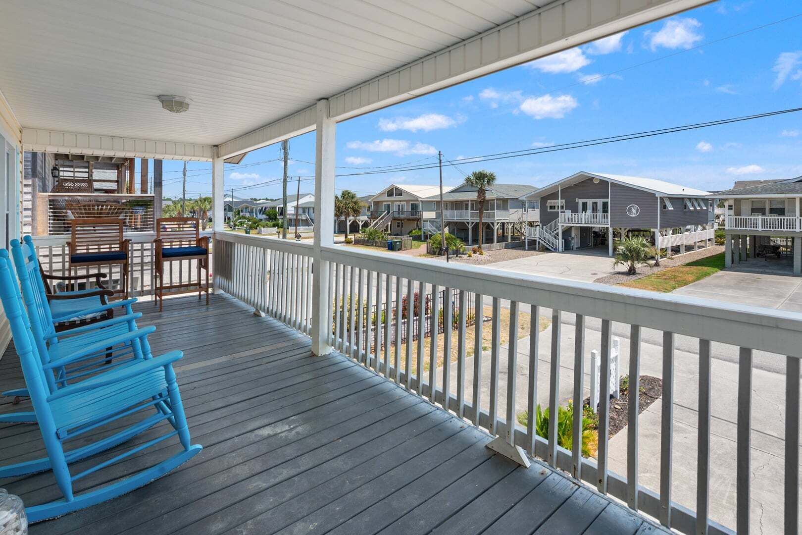A covered porch with blue rocking chairs, a white railing, and a view of a residential area with houses and a clear sky.