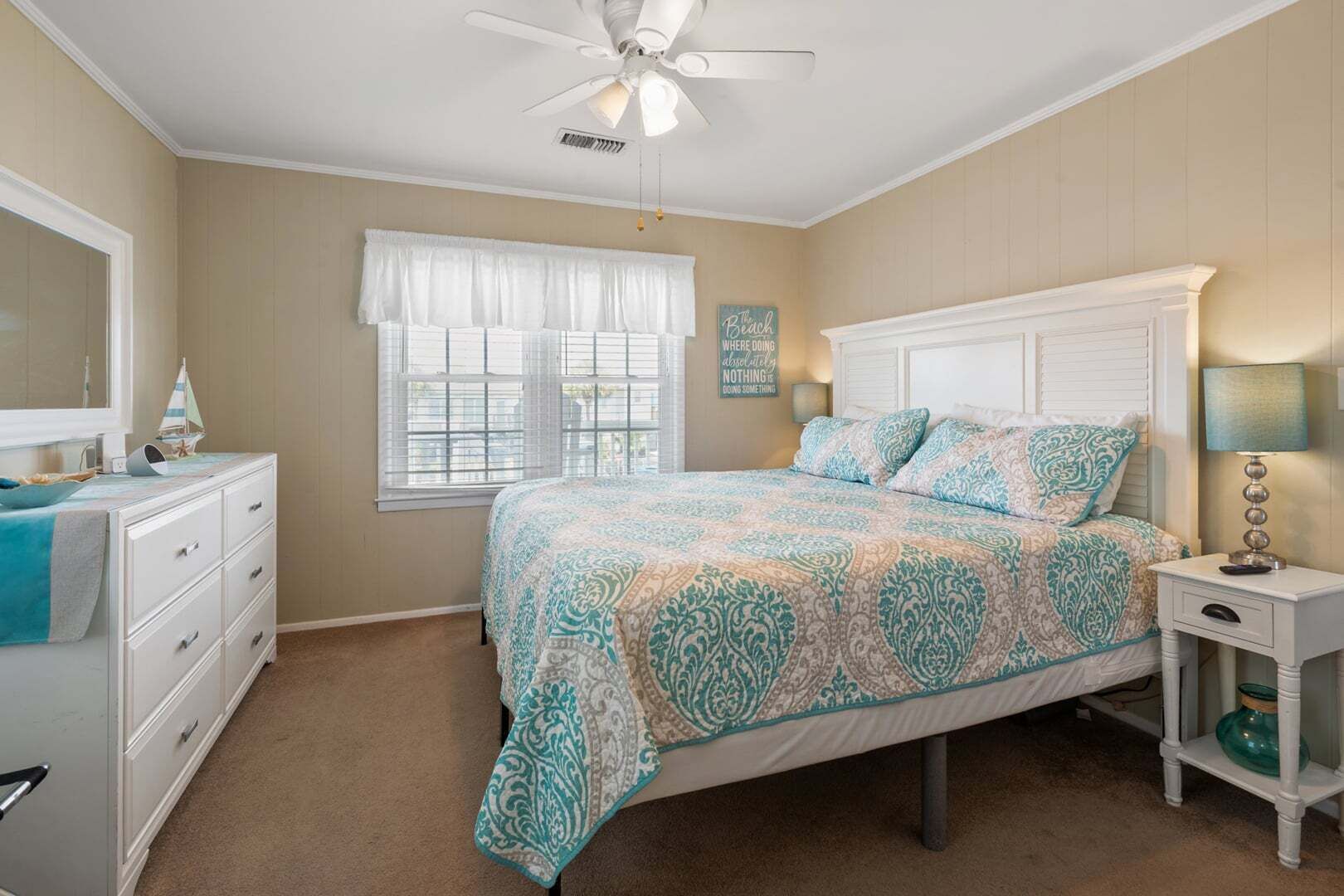 A bedroom with a bed, a nightstand, a dresser, a window with curtains, a ceiling fan, and a wall decoration.