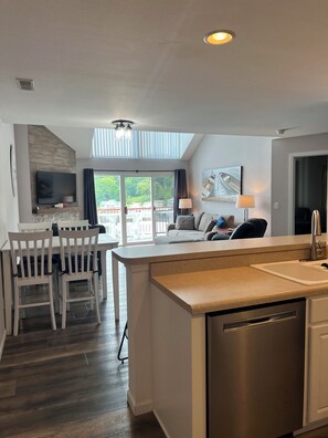 A modern kitchen with a built-in dishwasher, a dining area with white chairs, a large TV mounted on the wall, and a view of a dock and boats through the windows.