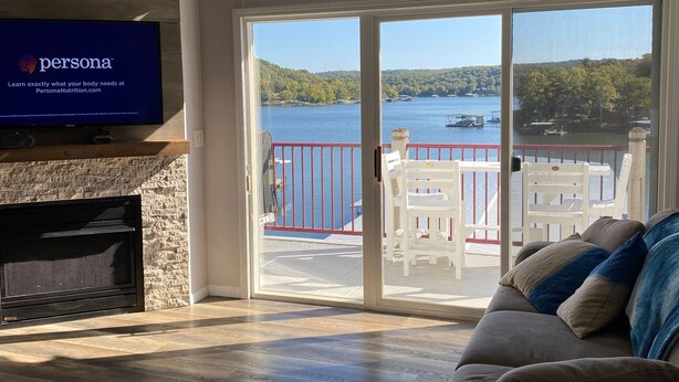 A modern living room with a fireplace, a television displaying 'Persona Nutrition', and a large sliding glass door leading to a balcony with a lake view. - 4BR/3BA Lakefront Penthouse, Clearwater Condominiums<br>Sleeps 8 (Camdenton)