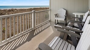 Een balkon met uitzicht op het strand, met een witte railing, twee Adirondack-stoelen en een kleine ronde tafel.