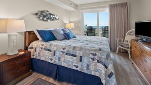 Een slaapkamer met een groot bed, een televisie, een kaptafel en uitzicht op het strand door het raam.