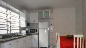 Cozinha com armários brancos, micro-ondas e geladeira. Há uma mesa de jantar com toalha vermelha e uma cadeira.
