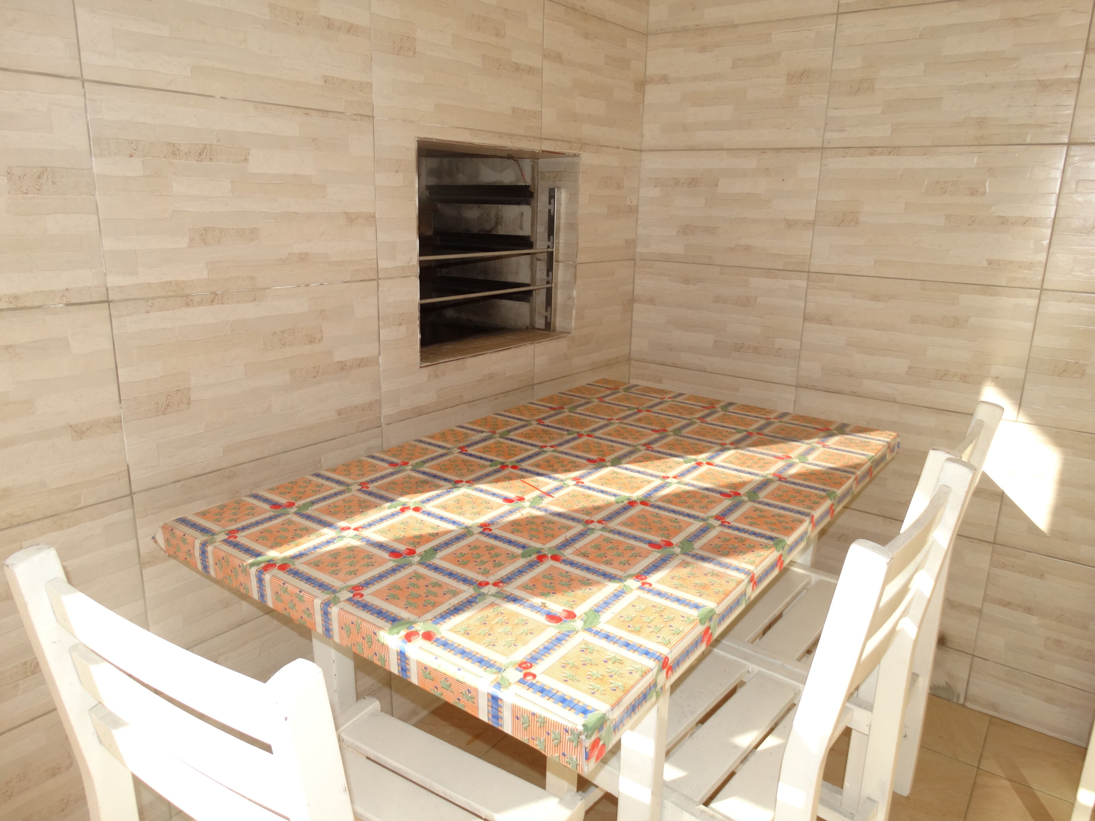 Une pièce avec un mur recouvert de carreaux, un four encastré, une table recouverte d’une nappe à motifs et des chaises blanches.