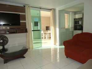 A modern living room with a flat-screen TV, a glass-top coffee table, a red sofa, and a sliding glass door leading to a balcony. - House 200m From the Sea - 9 (Florianópolis)