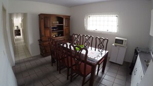A dining area with a wooden table and chairs, a kitchen with a microwave and a small refrigerator, and a window with blinds. - House 4D, 100m From the Sea - 229 (Florianópolis)
