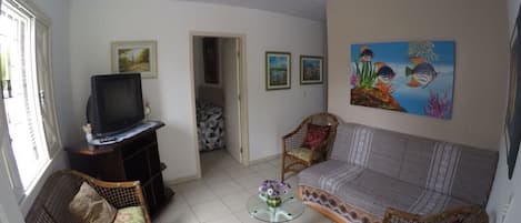 A living room with a sofa, two wicker chairs, a glass coffee table, a television on a stand, and framed pictures on the walls.