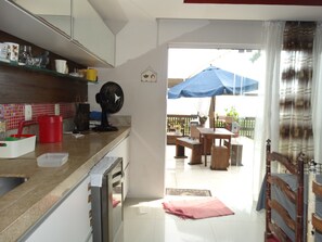 Cozinha equipada com geladeira, pia e ventilador. Possui área de refeições com mesa e cadeiras, e um guarda-chuva azul.
