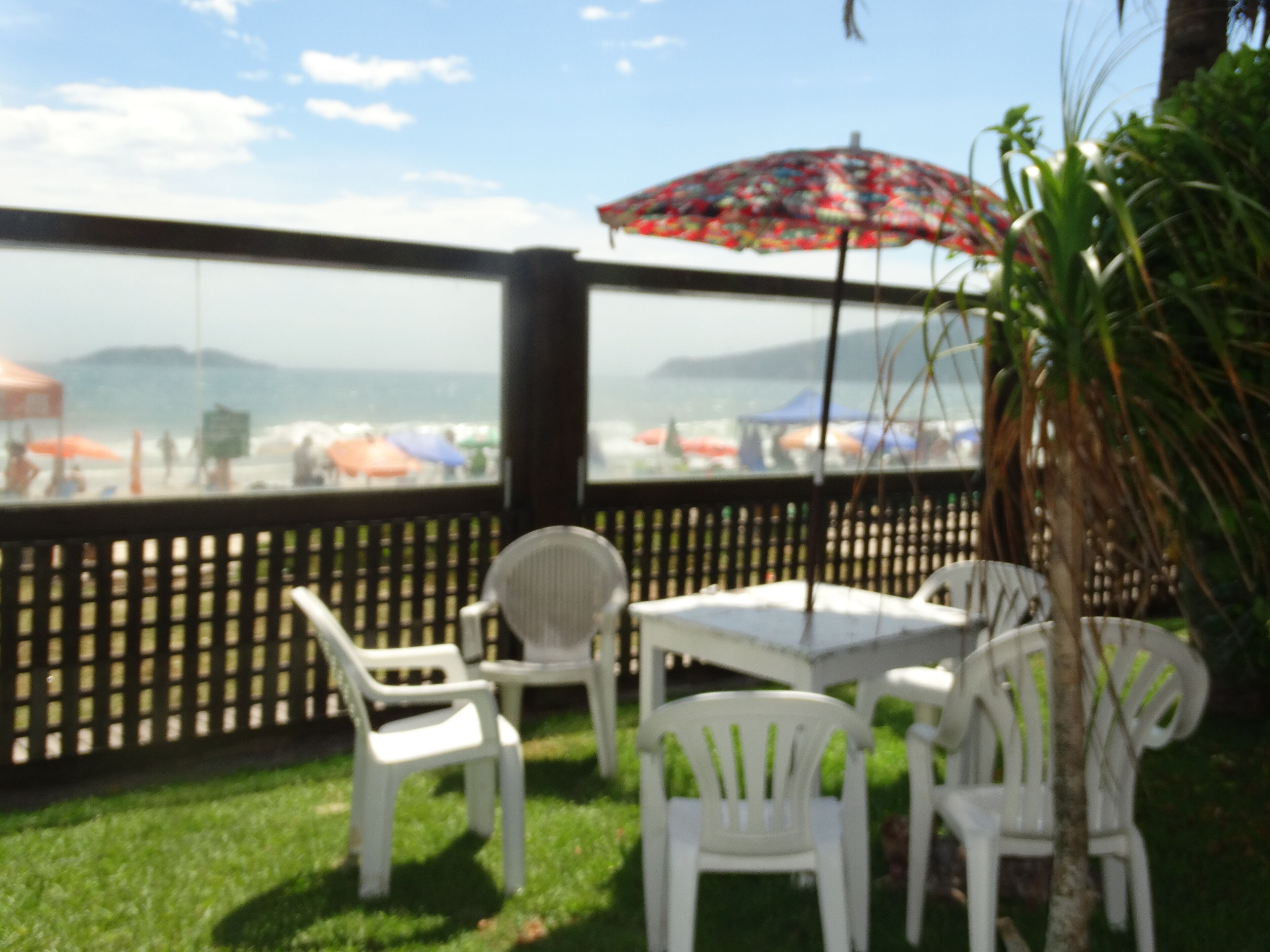 A patio area with white plastic chairs and a table, overlooking a beach with umbrellas.