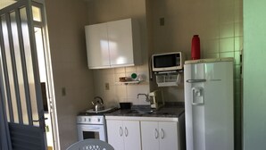 A compact kitchen with white cabinets, a microwave, a stove, and a refrigerator. There is a dining table with a chair and a white trash bin. - Apartment 1D, 50m From the Sea - 65 (Florianópolis)