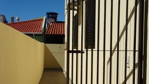 A narrow alleyway with a yellow wall on one side and a white wall with black metal bars on the other. There is a red-tiled roof visible in the background. - Apartment 1D, 50m From the Sea - 65 (Florianópolis)