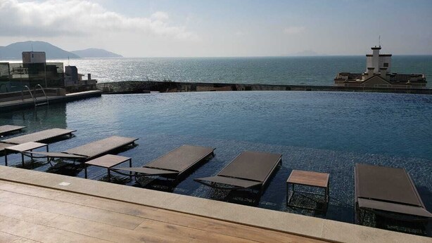 An infinity pool with lounge chairs, a wooden deck, and a view of the sea and distant mountains. - Apartment with Infinity Pool & Panoramic Views - 228 (Florianópolis)