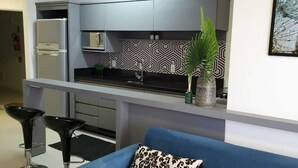 A compact kitchen with a refrigerator, microwave, and a patterned backsplash. There is a blue sofa with a decorative pillow and a plant on the counter. - Apartment with Infinity Pool & Panoramic Views - 228 (Florianópolis)