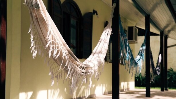 A covered outdoor area with a hammock hanging between columns, a tiled floor, and a building with arched windows.