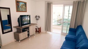 A living room with a blue sofa, a TV on a wooden stand, a fan, and a balcony with curtains. - 2 Bedroom Apartment - 202 (Florianópolis)