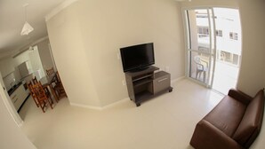 Ein Wohnzimmer mit einem braunen Sofa, einem Fernseher auf einem Schrank, einem Essbereich mit Holzsesseln und einem Tisch sowie einem großen Fenster mit Blick auf Gebäude.