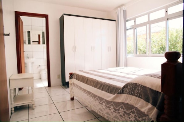 A bedroom with a bed, a wardrobe, a small table, and a window with a view of greenery.