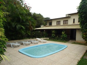 Una piscina con sillas de descanso, rodeada de exuberante vegetación y un edificio con tejado de tejas.