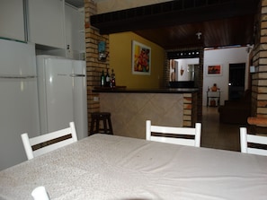 A kitchen with a dining area, a refrigerator, a counter with bottles, and a painting on the wall. - 4 Bedroom Pool House - 50 (Florianópolis)